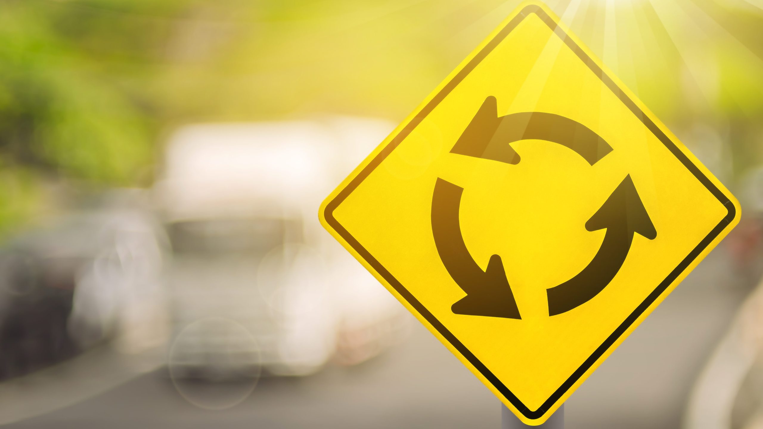 Roundabout warning sign on blur traffic road with colorful bokeh light abstract background. Copy space of transportation and travel concept. Retro tone filter color style. (Adobe Stock)