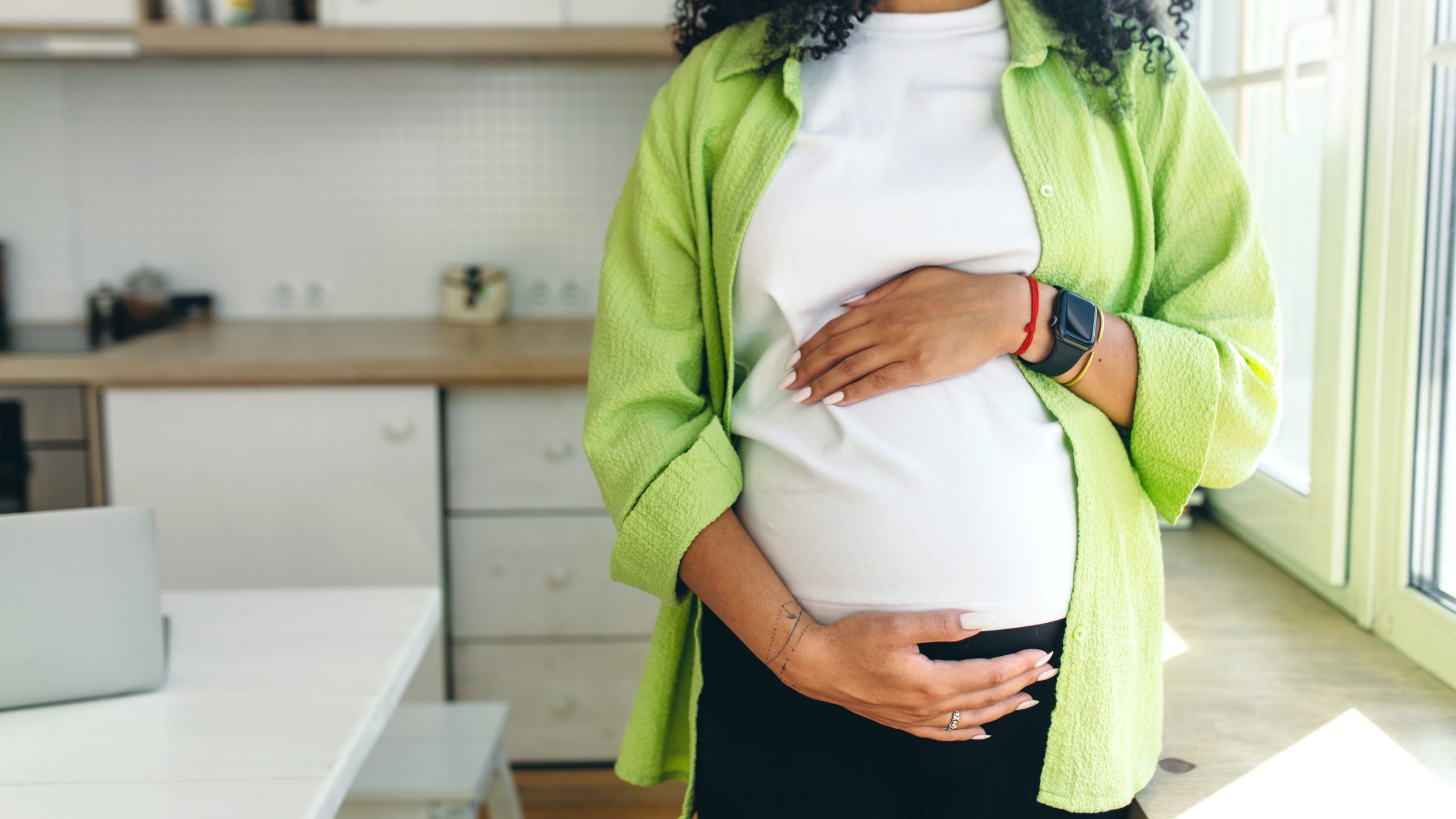 picture of pregnant african American woman with long curly hair looking through window standing in kitchen, rubbing her big cute belly, expecting baby boy to be born in few month