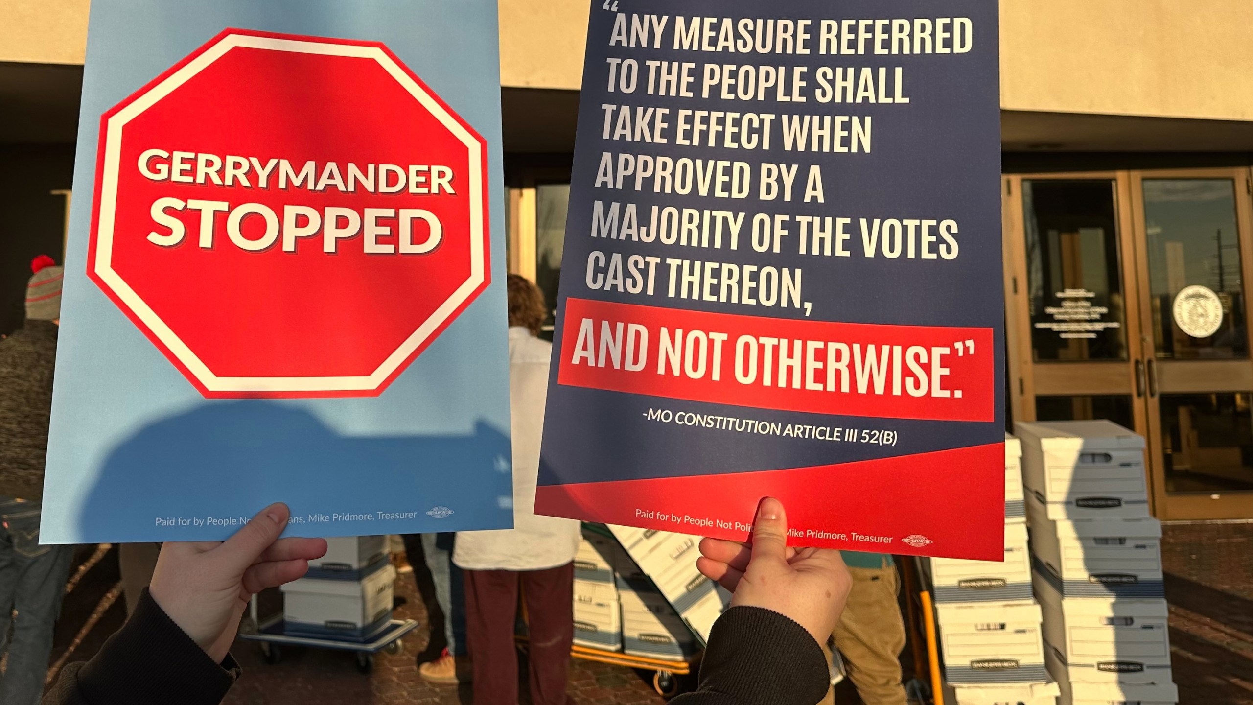 An opponent of new Missouri congressional districts approved by the Republican-led legislature holds signs outside the secretary of state's office in Jefferson City, Missouri, on Tuesday, Dec. 9, 2025, as petitions calling for a referendum election are delivered. (AP Photo/David A. Lieb)
